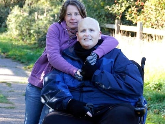 an woman standing and having her hand around a man who is sitting a wheels chair in a garden